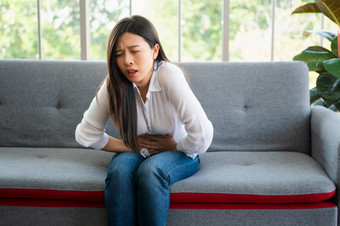 不开心亚洲女人坐着的沙发和持有胃痛苦腹部疼痛那来了从<strong>月经</strong>腹泻消化不良病和医疗保健概念