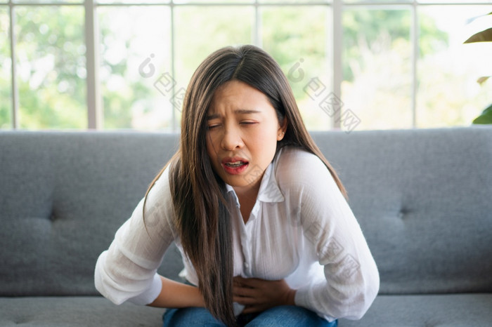 不开心亚洲女人坐着的沙发和持有胃痛苦腹部疼痛那来了从月经腹泻消化不良病和医疗保健概念