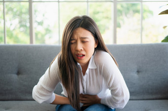 不开心亚洲女人坐着的沙发和持有胃痛苦腹部疼痛那来了从月经<strong>腹泻</strong>消化不良病和医疗保健概念