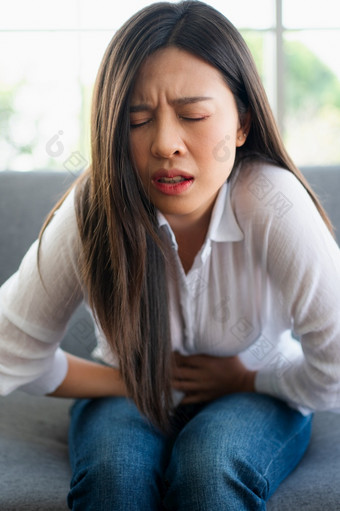 不开心亚洲<strong>女人坐着的</strong>沙发和持有胃痛苦腹部疼痛那来了从月经腹泻消化不良病和医疗保健概念