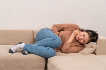 不开心亚洲女人坐着的沙发和持有胃痛苦腹部疼痛那来了从月经腹泻<strong>消化不良</strong>病和医疗保健概念