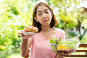 的快乐美丽的亚洲健康的中间岁的女人坐着的阳台旁边的花园和选择之间的甜甜圈和蔬菜沙拉概念健康哪和有营养的食物