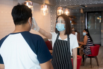 亚洲女服务员女人穿脸面具和持有红外额头温度计检查身体温度为病毒症状客户之前进入的餐厅咖啡商店