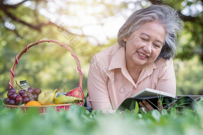快乐亚洲老高级女人和穿健康看和阅读书和说谎的野餐席公园和篮子水果除了概念快乐上了年纪的女人后退休和好健康