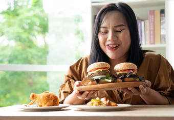 饿了超重女人持有汉堡木板炸鸡和披萨表格在工作从首页获得重量问题概念暴吃障碍床上