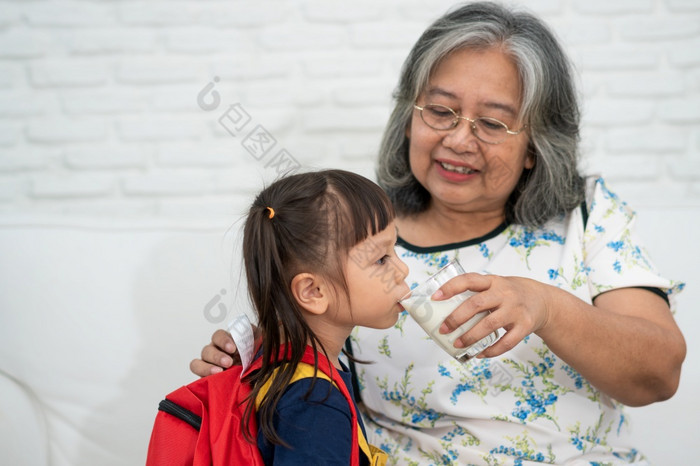 快乐上了年纪的亚洲奶奶坐在旁边她的（外）孙女和提要新鲜的牛奶从玻璃为早餐首页概念快乐家庭和需要哪在一起学前教育健康哪