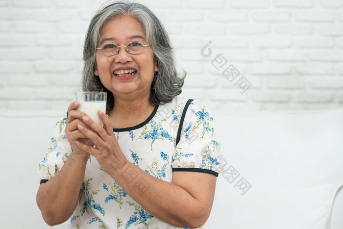 亚洲高级女人持有玻璃牛奶而放松沙发生活房间为退休健康上了年纪的女人喝玻璃牛奶维护她的幸福概念健康和健康的