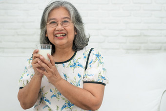 亚洲高级女人持有玻璃牛奶而放松沙发生活房间为退休健康上了年纪的女人喝玻璃牛奶维护她的幸福概念健康和健康的