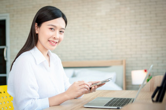 快乐美丽的亚洲女人工作移动PC和智能手机的首页办公室坐着表格快乐女专业自由职业者在线使用笔记本和智能手机概念