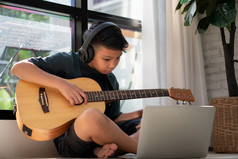 亚洲男孩玩吉他和看在线课程移动PC而练习为学习音乐音乐的仪器在线首页男孩学生研究在线与视频调用老师玩吉他