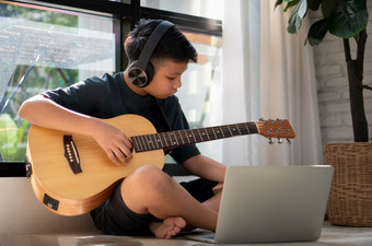 亚洲<strong>男</strong>孩玩吉他和看在线课程移动PC而练习为学习音乐音乐的仪器在线首页<strong>男</strong>孩学生研究在线与视频调用<strong>老师</strong>玩吉他