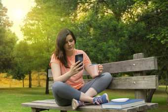 美丽的年轻的拉美裔女人坐着木板凳上的公园穿着运动服装与杯咖啡她的手阅读消息她的智能手机与绿色树背景美丽的年轻的拉美裔女人坐着木板凳上的公园与杯咖啡她的手阅读消息她的智能手机