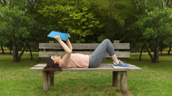 美丽的拉美裔年轻的女人运动服装说谎的木板凳上的中间公园没有人阅读蓝色的书对背景绿色树年轻的女人运动服装说谎的木板凳上阅读书