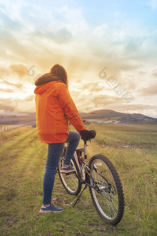 年轻的拉美裔女人穿橙色夹克和蓝色的裤子休息倾斜她的自行车见过从后面的中间的农村场在日落年轻的拉美裔女人穿橙色夹克和蓝色的裤子休息倾斜她的自行车的中间农村场在日落