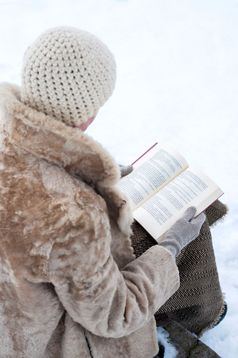 成人女人与皮毛外套和羊毛帽坐着石头板凳上和阅读书冬天公园覆盖雪