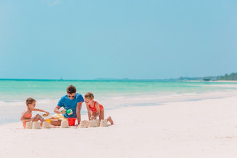 父亲和两个女孩玩与沙子热带海滩父亲和孩子们使沙子城堡热带海滩家庭玩与海滩玩具