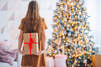 美丽的小女孩与礼物回来视图孩子持有礼物盒子附近的圣诞节树在室内快乐圣诞节和快乐假期回来视图孩子持有礼物盒子附近的圣诞节树在室内