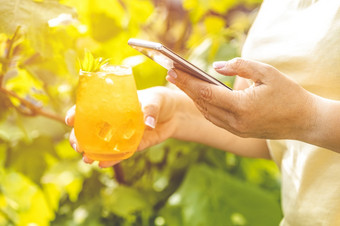 女人手使用移动电话和持有让人耳目一新橙色喝与冰夏天阳光明媚的花园背景技术我们的生活夏天时间
