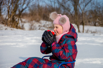 可爱的女孩与红色的杯坐在雪橇的雪的孩子变暖自己与热茶阳光明媚的冬天一天可爱的女孩与红色的杯坐在雪橇的雪的孩子变暖自己与热茶