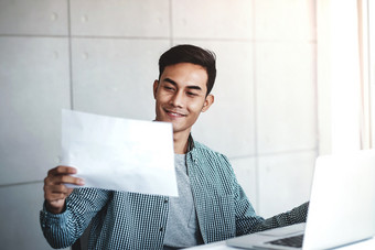 快乐年轻的亚洲商人工作电脑移动PC他的工作场所微笑和看纸工作表