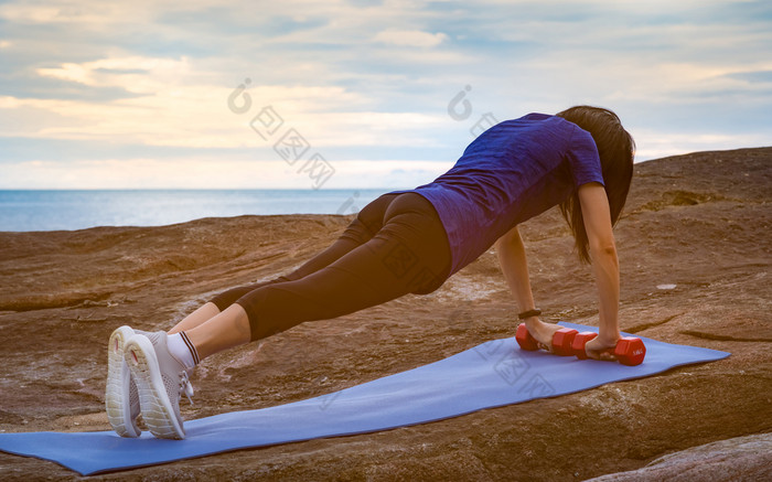 亚洲女人锻炼的早....石头海滩适合女人手持有红色的哑铃和推蓝色的瑜伽席为身体强度重量培训锻炼为健康的生活方式平屁股女人
