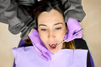 年轻的女病人参观牙医办公室美丽的女人坐着牙科椅子与开放口在口服检查而医生工作牙齿高质量照片年轻的女病人参观牙医办公室美丽的女人坐着牙科椅子与开放口在口服检查而医生工作牙齿