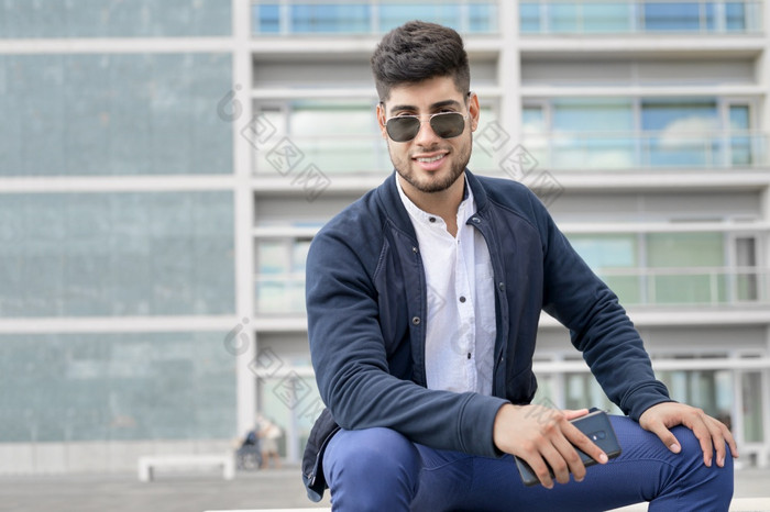 肖像年轻的男人。时尚的太阳镜摆姿势而坐着板凳上城市背景高质量照片肖像年轻的男人。时尚的太阳镜摆姿势而坐着板凳上城市背景