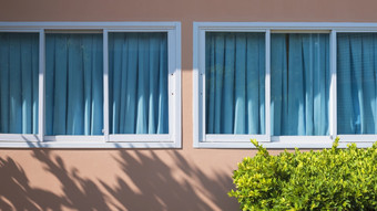玻璃滑动窗户和蓝色的窗帘粉红色的水泥墙与阳光和影子表面和绿色布什前景首页外体系结构概念