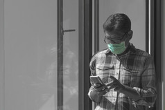 亚洲男人。穿保护面具和使用智能手机附近玻璃窗口生活房间首页黑色的和白色与飞溅绿色颜色技术