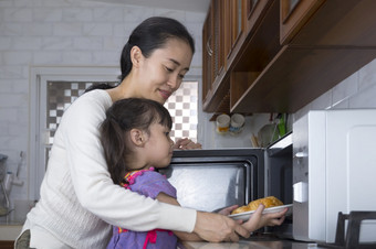 亚洲妈妈。教学她的小女儿使用微波烤箱温暖的羊角面包的厨房首页
