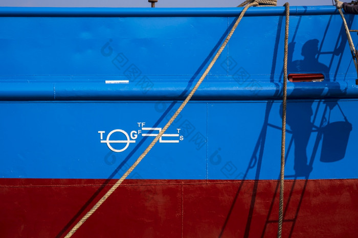 阳光和影子画家攀爬梯与白色橡胶底帆布鞋马克蓝色的和红色的生锈证明钢船体表面钓鱼船与系泊绳子船厂区域