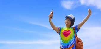 亚洲女旅行者色彩斑斓的休闲风<strong>格</strong>使用智能手机采取自拍与微笑<strong>高高</strong>兴兴地和提<strong>高</strong>她的手对白色云和蓝色的天空<strong>背景</strong>享受周末假期