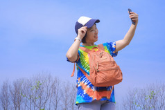 亚洲女旅游色彩斑斓的休闲风格微笑高高兴兴地和使用智能手机采取自拍与模糊无叶的森林蓝色的天空背景