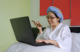快乐亚洲中间岁的女人白色<strong>浴</strong>袍与淋<strong>浴帽</strong>会说话的智能手机和使用移动PC她的床上早....时间