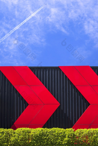 红色的箭头模式黑色的波纹金属墙与绿色布什栅栏公共健康公园对飞机飞机飞行蓝色的天空背景垂直框架