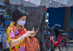 亚洲女旅行者色彩斑斓的休闲风格与保护面具使用智能手机人行道海边区域