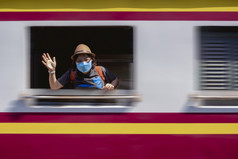 运动模糊女旅行者穿保护面具看相机和挥舞着手从火车窗口而的火车离开的站