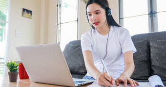 亚洲快乐女人学生穿耳机研究在线看网络研讨会播客移动PC听学习教育课程会议调用和写笔记看移动PC坐沙发首页