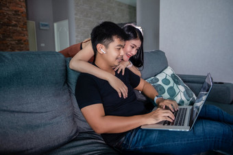 亚洲年轻的微笑夫妇拥抱和使用无线耳机和使用电脑笔记本工作从首页准备图和信息为在线会议首页
