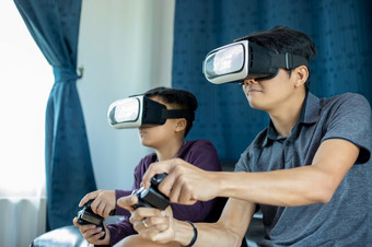 亚洲父亲和儿子享受玩视频游戏在一起与视频操纵杆和虚拟现实眼镜与令人兴奋的和非常有趣的的生活房间首页