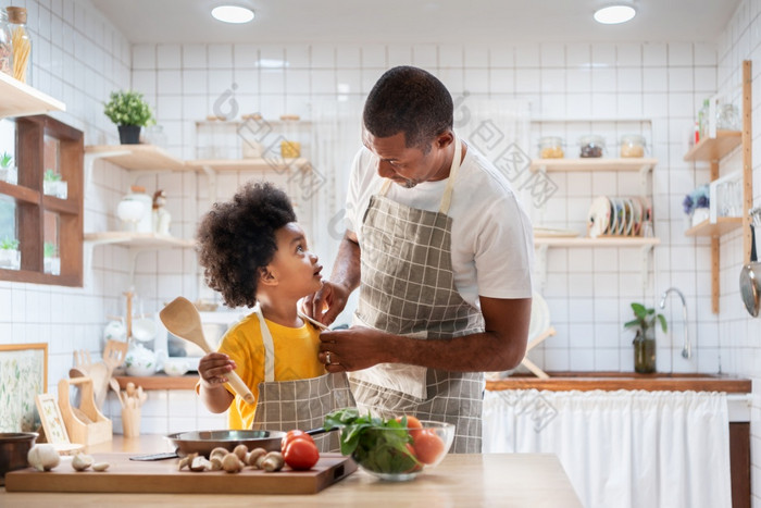 快乐非洲父亲和儿子衣服在一起之前烹饪的白色厨房单爸爸老板与黑色的孩子助手黄色的衬衫准备食物和看每一个其他首页眼睛联系的关系会说话的温暖的家庭