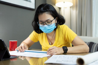 亚洲女架构师穿眼镜使用规模统治者为测量她的蓝图在检疫流感大流行冠状病毒年轻的女人室内设计师黄色的衬衫蓝色的外科手术面具工作从首页在科维德