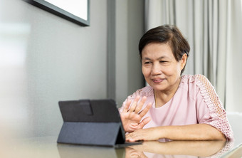 快乐微笑高级亚洲女人使<strong>视频</strong>调用数字平板电脑在检疫和孤立的首页上了年纪的女<strong>粉红</strong>色的布挥舞着手而会说话的与人通过互联网技术