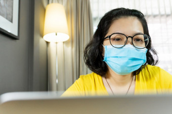 快乐亚洲女穿眼镜使用移动PC电脑在检疫孤立的年轻的女人黄色的衬衫穿蓝色的外科手术面具工作从首页在流感大流行病毒新冠病毒冠状病毒健康的预防保护