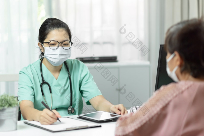 亚洲女医生绿色统一的穿眼镜和外科手术面具写作某物检查表文档而会说话的与上了年纪的女人病人医疗房间的医院症状疾病流感新冠病毒冠状病毒