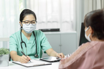 亚洲女医生绿色统一的穿眼镜和外科手术面具写作某物检查表文档而会说话的与上了年纪的女人病人医疗房间的医院症状疾病流感新冠病毒冠状病毒