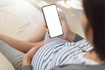 怀孕了女人使用智能手机和做在线购物沙发亚洲女享受使在线视频调用与移动电话在她的怀孕