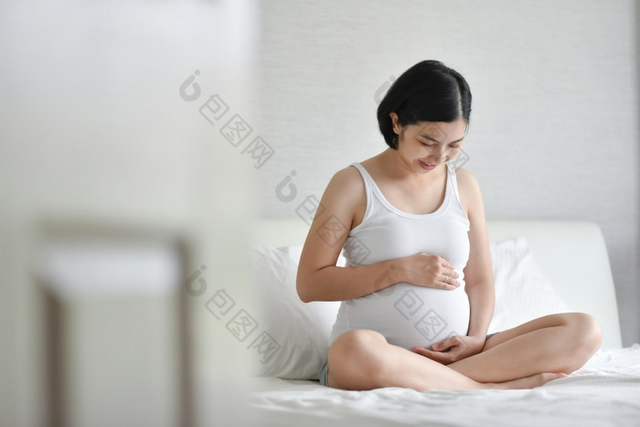 美丽的怀孕了亚洲女人白色睡衣微笑坐着床上健康的快乐年轻的妈妈。持有和触碰和看她的肚子首页而怀孕家庭孕妇生活方式