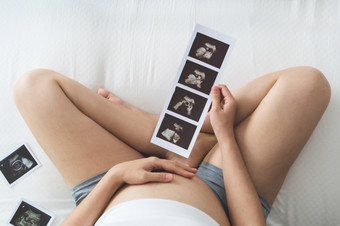 高角视图快乐亚洲怀孕了女人持有超声波图片而触摸她的肚子床上年轻的妈妈。与sonogram她的未<strong>出生</strong>的婴儿概念怀孕孕妇产前哪
