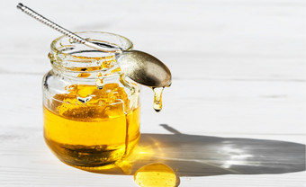 有机自然蜂蜜玻璃Jar白色木背景特写镜头古董勺子开放Jar蜂蜜蜂蜜滴的表格明亮的阳光照通过Jar蜂蜜